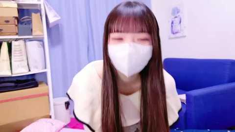 Media: Video of an Asian woman with long brown hair and bangs, wearing a white face mask and a light-colored top, sitting in a cluttered room with a blue sofa, shelves, and boxes in the background.