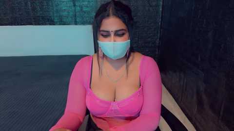 Media: Video of a plus-size woman with long dark hair, wearing a pink, long-sleeve, lace bralette and a face mask, seated on a mat in a dimly lit, black-walled room.