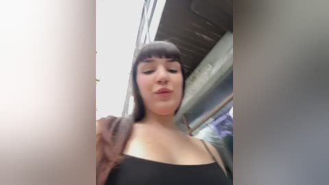 Media: Video of a young woman with straight black hair and bangs, wearing a black tank top and brown jacket, standing in a bus.