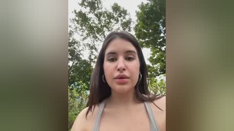 Media: Video of a young woman with light skin, long dark hair, and a neutral expression, wearing a gray tank top. Background features green foliage and a cloudy sky.