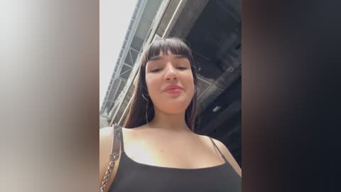 Media: Video of a young woman with long, straight brown hair and bangs, wearing a black tank top, standing in an industrial setting with metal beams and concrete.