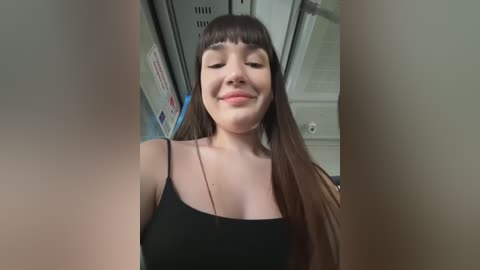 Media: Video of a smiling young woman with long brown hair and bangs, wearing a black spaghetti-strap top, inside a dimly lit elevator.