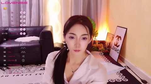 Media: Video of an Asian woman with long black hair, wearing a white blouse and black jacket, in a cozy living room with a TV and candles.