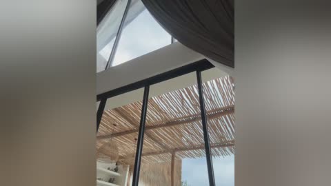 Media: Video of a modern, minimalist indoor space featuring a large, triangular skylight and a wooden pergola with bamboo slats. The room has a clean, white wall and a partial view of a tropical, beach-like exterior.