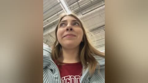 Media: Video of a smiling young woman with light skin, shoulder-length brown hair, wearing a red T-shirt with white text and a blue-striped hoodie, standing in a brick-lined hallway with exposed ceiling beams.