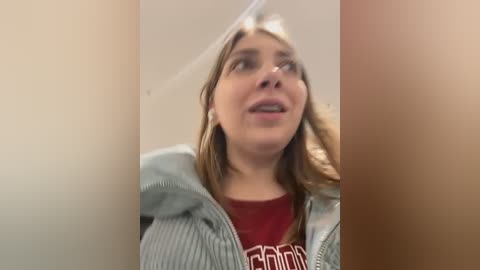 Media: Video of a young woman with light skin, light brown hair, wearing a red t-shirt and a gray hoodie, looking up, slightly out of focus, against a beige wall.