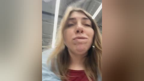 Media: Video of a young woman with long, blonde hair, wearing a red top, puckering her lips in a selfie. Background shows a dimly lit, industrial space with exposed beams and fluorescent lights.