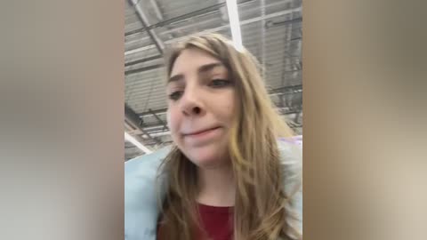 Media: Video of a young Caucasian woman with long, wavy light brown hair, wearing a red top and light blue jacket. She smiles slightly in an indoor, industrial setting with a metal ceiling.