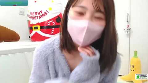 Media: Video of an Asian woman with straight, shoulder-length brown hair, wearing a light blue fuzzy robe and a pink face mask, smiling, in a bright, white kitchen with a \"Merry Christmas\" red Santa sack and a yellow banana toy.