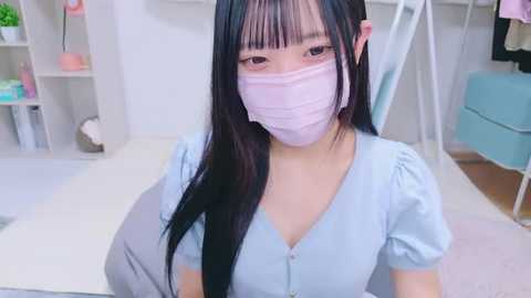 Media: Video of an Asian woman with long black hair, wearing a pink face mask, light blue shirt, and sitting on a white floor in a modern, minimalist room.