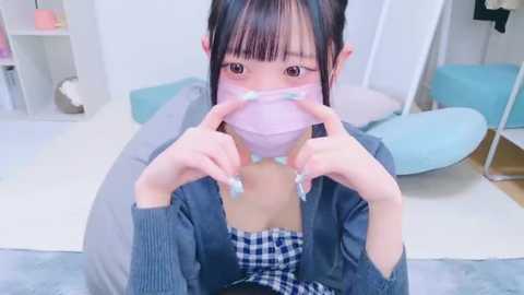 Media: Video of a young Asian woman with straight black hair, wearing a pink face mask and blue checkered top, holding her mask, in a modern, light-colored bedroom with blue furniture.