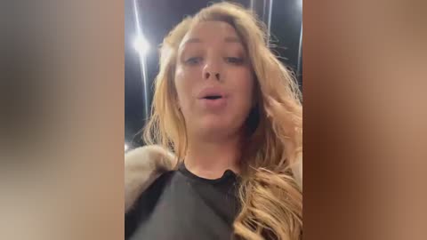 Media: Video of a blonde woman with wavy hair, wearing a black sheer top, slightly parted lips, and a neutral expression, taken indoors with blurred background.