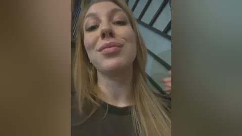 Media: Video of a smiling, fair-skinned, blonde woman with long hair, wearing a black top, standing indoors near a metal staircase.