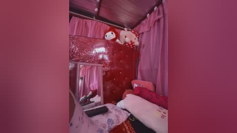 Media: Video of a small, cozy bedroom with a red quilt, stuffed animal, pink curtains, and a mirror on the wall, all bathed in pink lighting, giving a warm, intimate atmosphere.