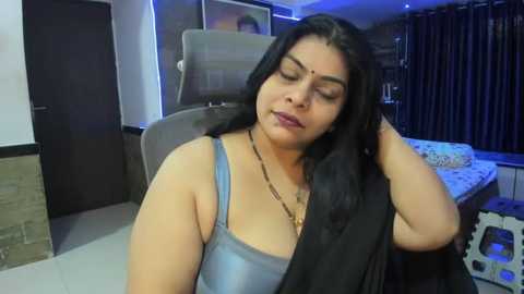 Media: Video of a middle-aged Indian woman with long black hair, wearing a sleeveless blue top, sitting in a modern living room with dark curtains and white furniture.