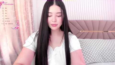 Media: Video of an Asian woman with long black hair, wearing a white blouse, sitting on a bed with patterned pillows and a tufted headboard. The room has pastel-colored curtains and a soft, feminine ambiance.