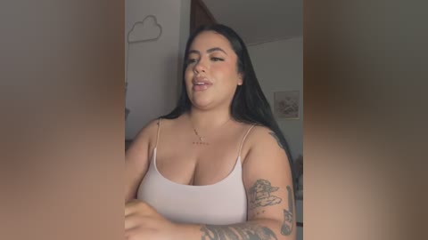 Media: Video of a plus-size Latina woman with long black hair, wearing a white tank top, standing in a dimly lit room with a cloud-shaped wall decal and framed art.