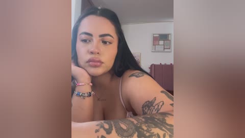 Media: Video of a young woman with long black hair, light skin, and a tattoo sleeve, wearing a spaghetti-strap top, sitting on a bed with a red bedspread, in a room with a framed abstract painting on the wall.