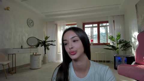 Media: Video of a young East Asian woman with long black hair, wearing a light blue shirt, in a minimalist, white-walled room with plants, a round mirror, and a clock.