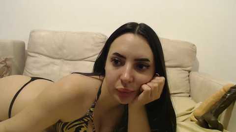 Media: Video of a young woman with long black hair, light skin, and medium-sized breasts, wearing a black and yellow zebra-print bikini, lying on a beige sofa, resting her head on her hand.