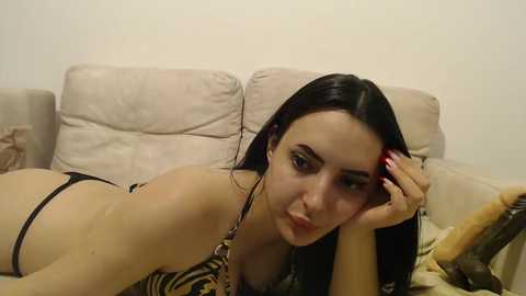 Media: Video of a young woman with fair skin, long black hair, lying on a beige couch, wearing a leopard-print bikini. She rests her head on her hand, gazing directly at the camera.