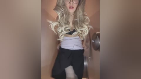 Video of a young woman with long, wavy blonde hair, wearing glasses, a black bra, and black pants, kneeling in a dimly lit, beige hallway.