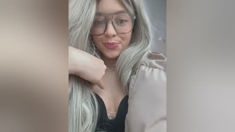 Video of a fair-skinned woman with long, platinum blonde hair and large, round glasses, wearing a black lace bra and beige jacket, resting her hand on her chin.