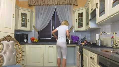 Media: Video of a blonde woman in white leggings and a short-sleeved top, cooking in a cream-colored kitchen with white cabinets, a window with sheer curtains, and a silver sink.