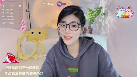 Media: A video of an Asian woman with black hair, wearing glasses and a gray hoodie, smiling. The background features a yellow plush toy and a potted plant.