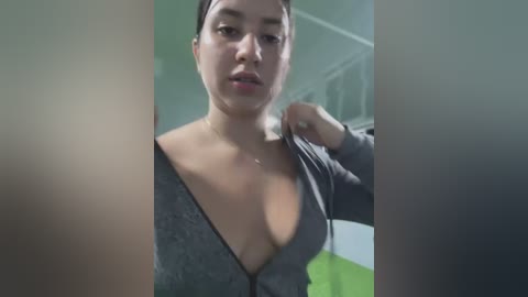 Media: Video of a Latina woman with medium skin tone and dark hair, wearing a gray zipper hoodie, standing in a dimly lit room with green and white walls.