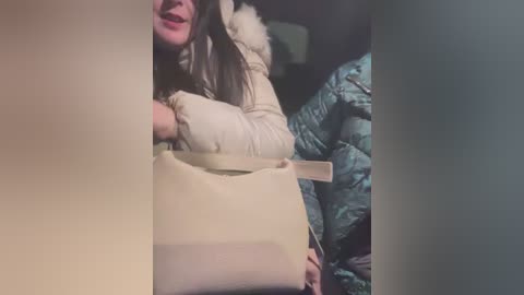 Media: A video captures a woman with long, dark hair and a beige jacket, carrying a beige handbag, sitting on a teal, puffy blanket in a dimly lit room.