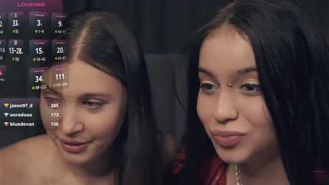 Media: Video of two young women with fair skin and long black hair, one with a nose ring, sitting close together, smiling, in a dimly-lit room.