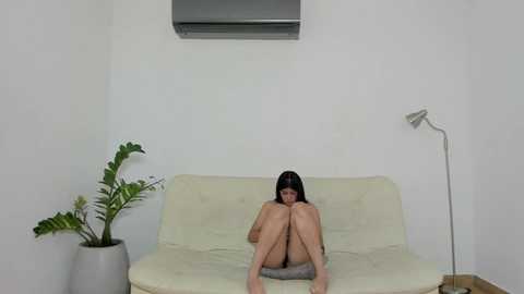 Media: Video of a naked woman with long black hair sitting on a white couch, covering her face with her knees, in a minimalist room with a potted plant and a floor lamp.