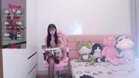 Media: Video of a young Asian woman in a pink gaming chair, wearing a pink top and black stockings, playing a video game on a white console. Pink teddy bears decorate the bed.