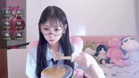 Media: A video of an East Asian woman with long black hair, glasses, and a light blue shirt, eating noodles on a pink couch with stuffed animals, in a bright room.