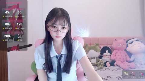 Media: A video of a young East Asian woman with long black hair, glasses, and a blue shirt, sitting on a pink gaming chair in a bedroom with stuffed toys.