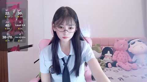 Media: A video of an East Asian woman with long black hair and glasses, wearing a white school uniform with a tie, sitting on a pink gaming chair in a bedroom with stuffed toys and a TV screen displaying game stats.