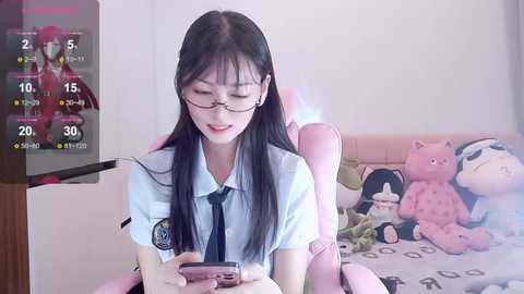 Media: Video of an East Asian woman with long black hair and glasses, dressed in a white school uniform, sitting in a pink gaming chair, surrounded by stuffed toys.