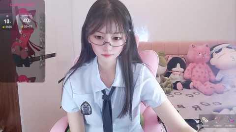 Media: Video of a young Asian woman with long black hair, glasses, and a white school uniform, sitting on a pink gaming chair in a room with plush toys, including a panda and a red cat.