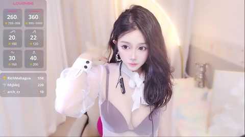 Media: A video of a young Asian woman with long black hair, fair skin, and a slender physique, wearing a sheer white top and a pink bra, standing in a softly lit room.