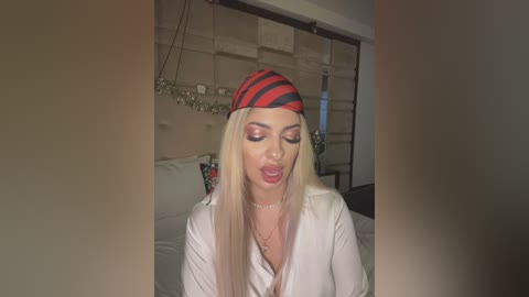 Media: A video of a blonde woman with long hair, wearing a red and black striped hat, a white shirt, and a necklace, posing seductively indoors with beige walls and a bed in the background.