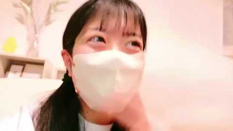 Media: A video of a young East Asian woman with long black hair, wearing a white face mask, looking upwards with a curious expression. The background features a pale wall, a wooden table, and a green plant.