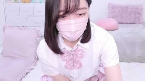 Media: Video of an East Asian woman with straight black hair and a light complexion, wearing a white blouse with a pink bow, pink mask, and pink ruffled skirt, kneeling on a pink and white bed. Soft pastel colors dominate the serene, tidy room.