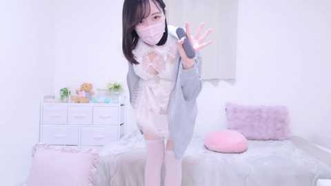 Media: Video of a slender, fair-skinned Asian woman with shoulder-length black hair, wearing a white lace dress, gray cardigan, and a face mask, standing in a minimalist, pastel-pink bedroom with plush pillows and a white dresser.