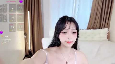 Media: A video of a young East Asian woman with long black hair, fair skin, and red lipstick, wearing a white spaghetti-strap top, sitting on a bed in a modern bedroom with beige curtains.