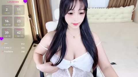 Media: A video of a young Asian woman with long black hair and fair skin, wearing a white lace lingerie top, sitting on a bed with beige pillows in a modern bedroom.