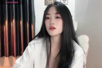 Media: A video of a young East Asian woman with long black hair, wearing a white robe, sitting in a modern, dimly-lit room with black curtains. She has a neutral expression.