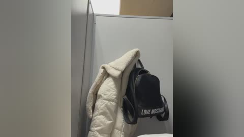 Media: A video of a beige quilted jacket with a black backpack featuring \"LOVE MOSCHINO\" text hanging on a plain white wall in a small, sparsely decorated room.