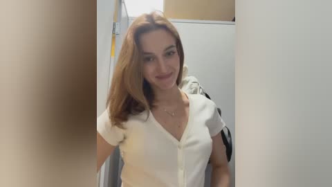 Media: Video of a young woman with long, light brown hair, wearing a white cardigan, standing in a dimly lit room with a white door and a beige wall in the background.