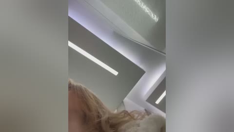 Media: Video of a woman with long, light brown hair, possibly in a white dress, standing in a modern, minimalist room with white walls and sleek, angular lighting fixtures.